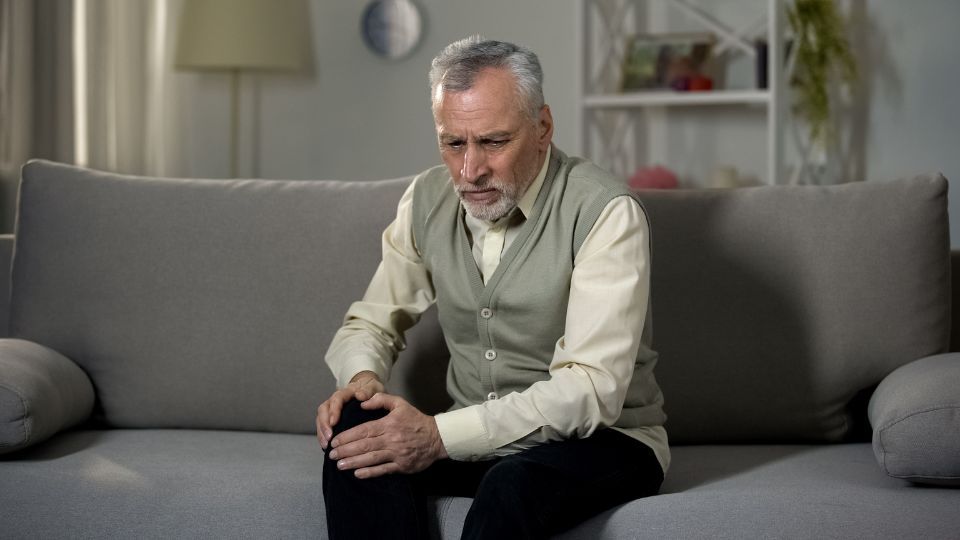 Elderly Man Holding His Knee In Pain On Couch